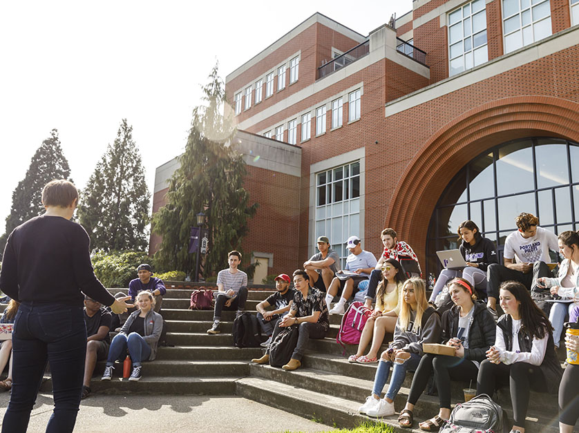 A group of prospective students sit on the steps and listen to a tour guide on University of Portland's campus