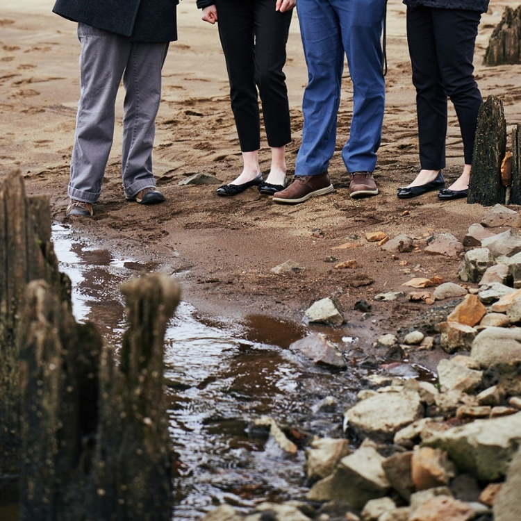 four people standing on muddy bank by driftwood and small water inlet.