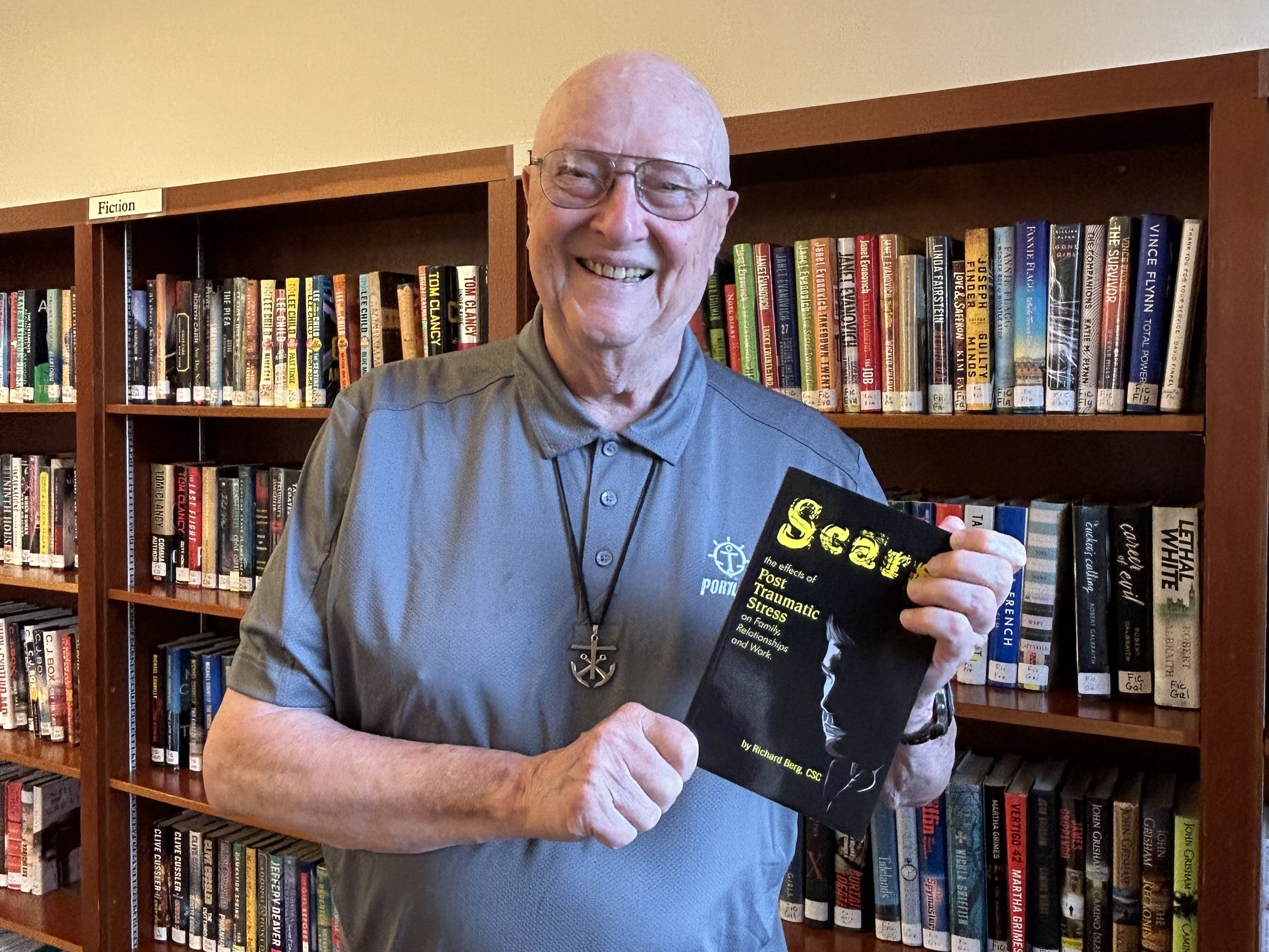 Father Berg stands smiling in a library holding a copy of his book "Scars"