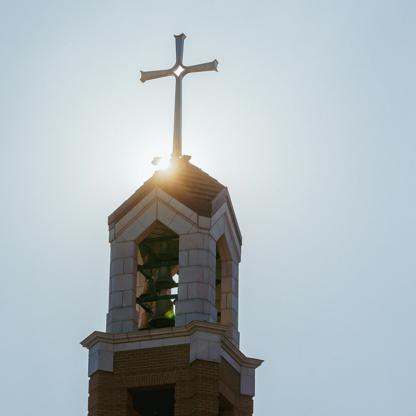 The top of University of Portland's chapel bell tower with a cross steeple backlit by sunlight against a clear blue sky.