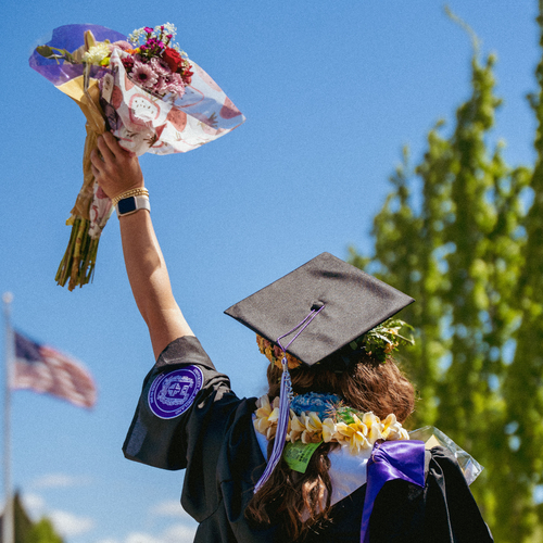 A behind view of a University of Portland graduate in a cap and gown lifting a bouquet of flowers high in the air.