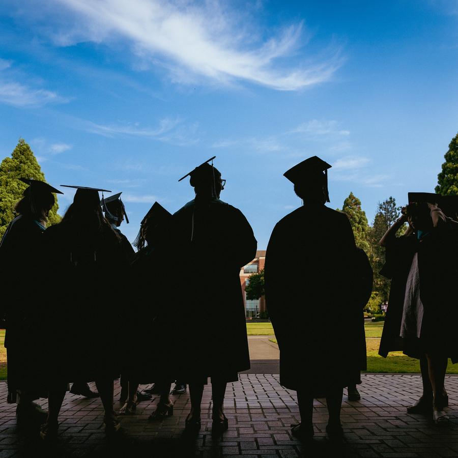A group of University of Portland graduates in caps and gowns, silhouetted against a bright blue sky.