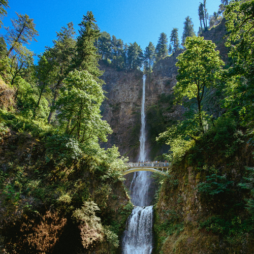 A tall waterfall cascades down a rocky cliff and University of Portland students stand on a small bridge in front of it.
