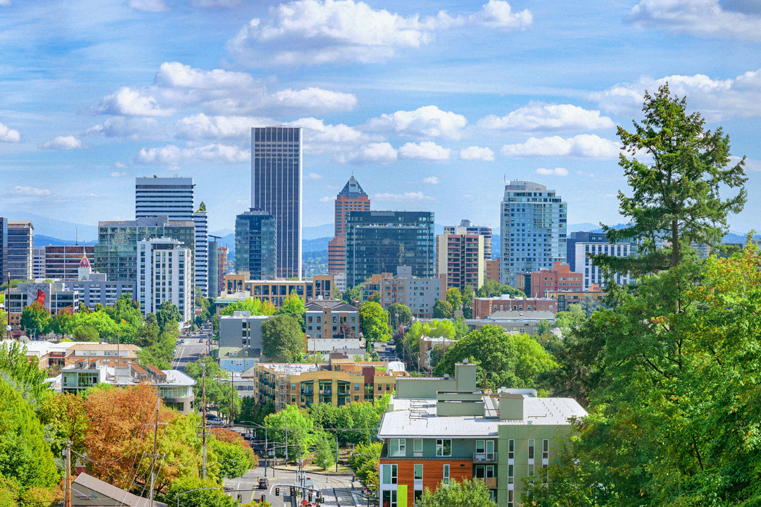 An aerial view of the skyline of downtown Portland, Oregon, with green trees and vibrant blue sky.