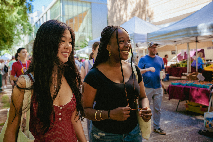 Two University of Portland students smile while walking through the outdoor Portland Farmers Market on a sunny day.