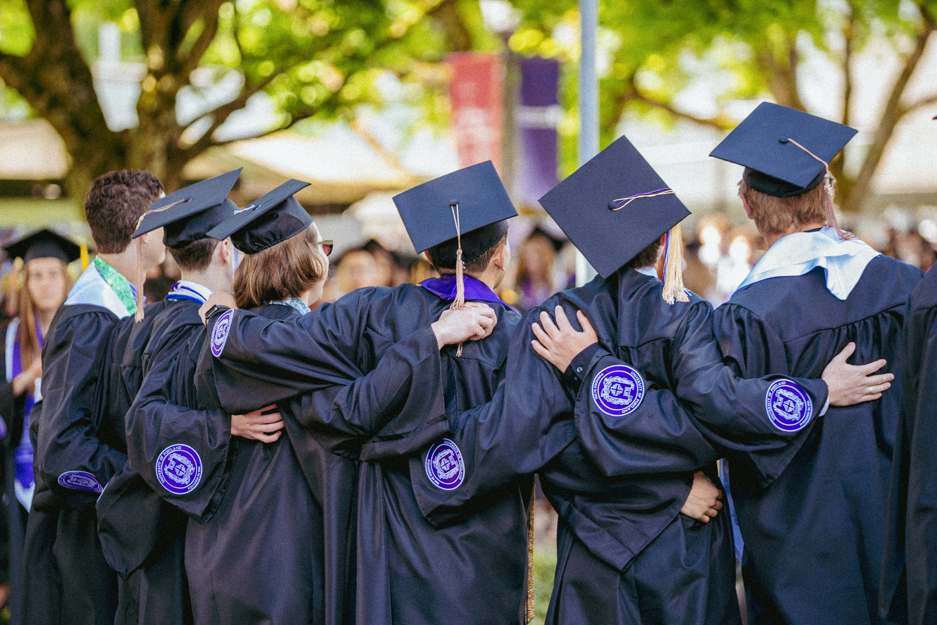A behind view of a group of University of Portland graduates in caps and gowns standing with their arms around each other.