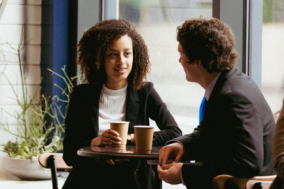 A UP student and professional colleague talk while sitting in a coffee shop, with brown cups on the table in front of them.