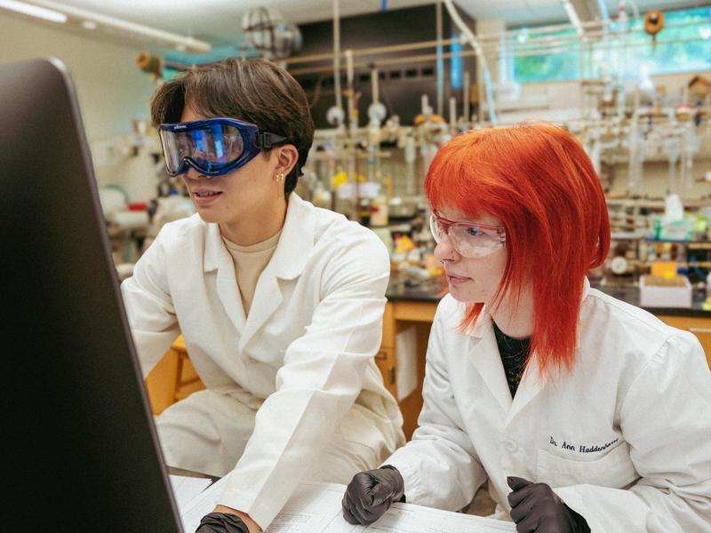 Two UP students wearing white lab coats and protective goggles look at a computer screen in a science lab.