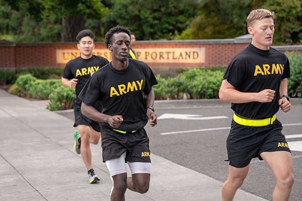 Rotc cadets running on campus past the entrance gate wearing army shirts 