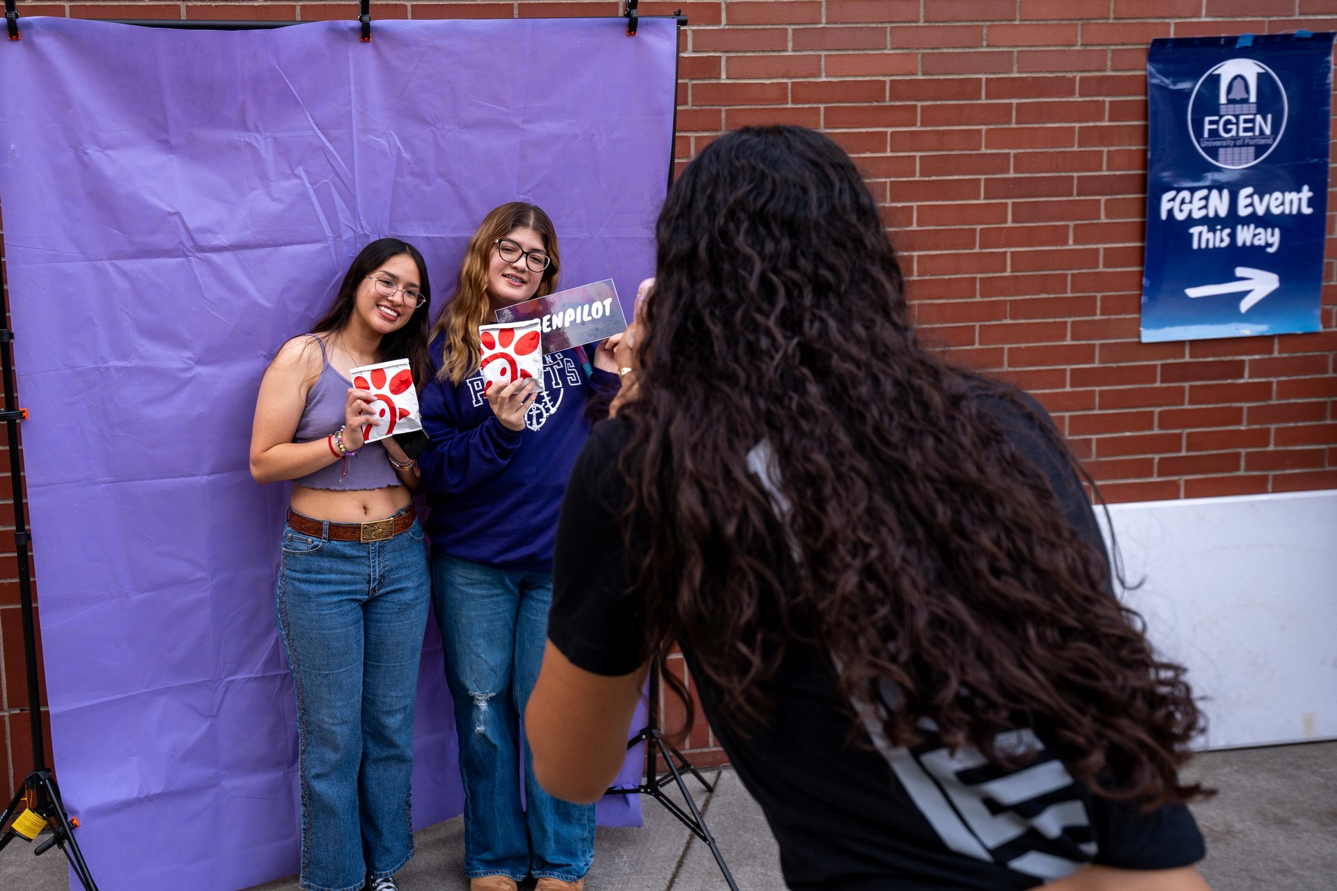 Two individuals pose with food and event signs in front of a purple backdrop, while a third person photographs them. A brick wall and an “FGEN Event This Way” sign are visible in the background.