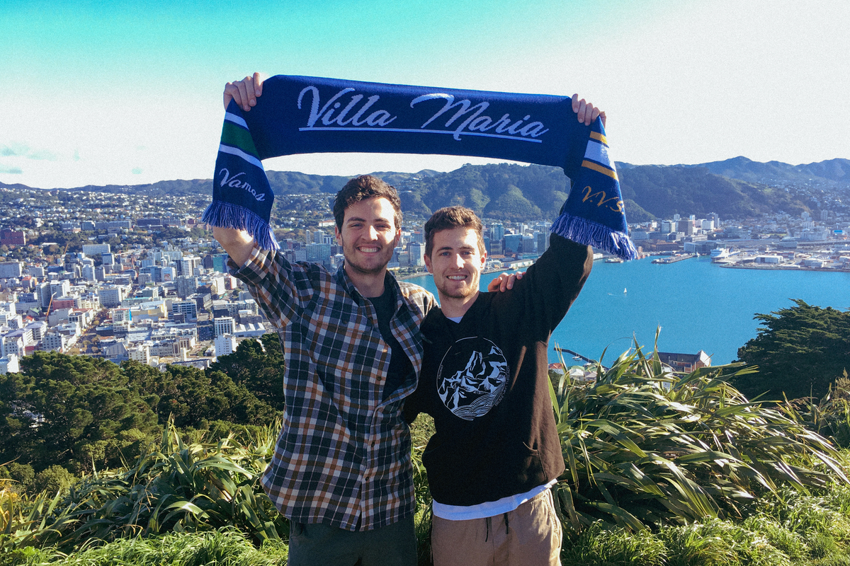 Two UP students stand on a ledge above New Zealand and hold up a scarf that says "Villa Maria" during a study abroad trip.