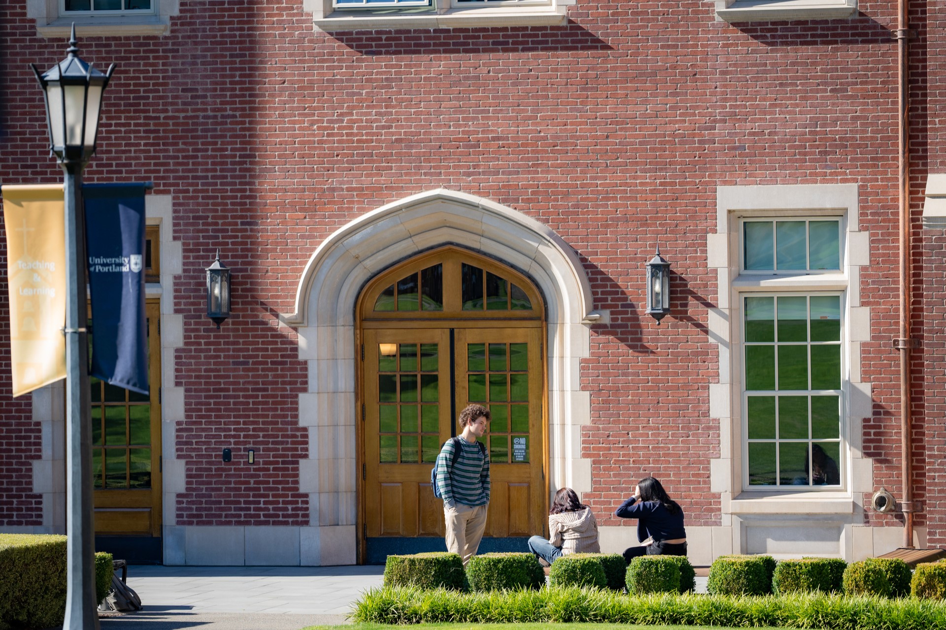 Three individuals converse outside a campus brick building featuring arched wooden doors and large windows. Two are seated on landscaped bushes, while one stands nearby. Purple and gold university banners are displayed on a lamppost.