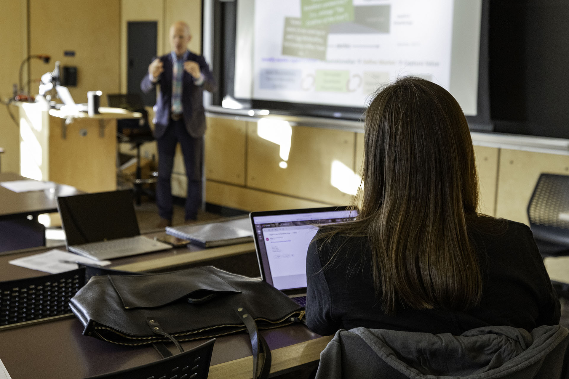 A graduate business student pictured from behind watches the professor lecture in front of the class