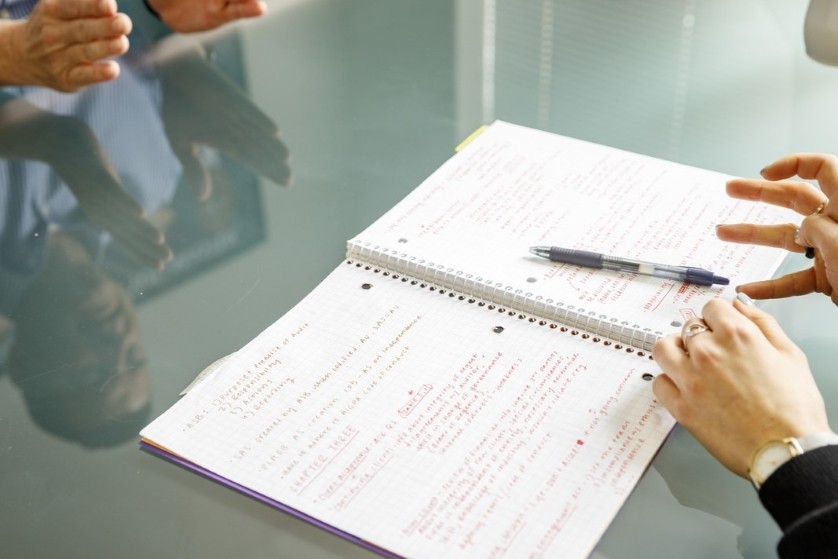A close up view of the hands of two people holding pens and working over a note pad.