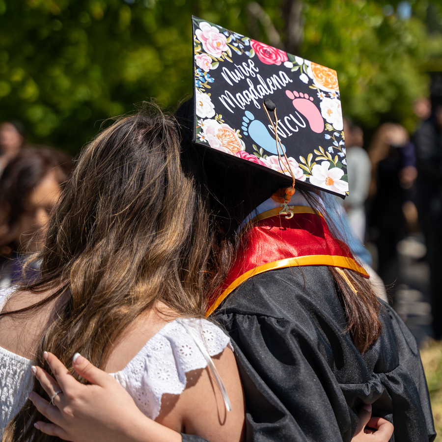 A student wearing a graduation cap adorned with flowers, baby footprints, and "Nurse Magdalena NICU" embraces a friend in an outdoor setting with greenery in background.
