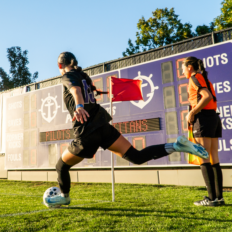 A Pilot women's soccer player with long hair in a ponytail and a black sports jersey stands in the corner of the field facing the scoreboard as she takes a corner kick. 
