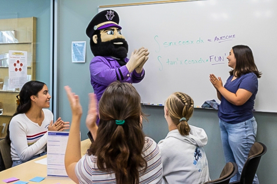 Wally Pilot, the University of Portland mascot, stands in front of a classroom whiteboard while engaging with students during a lecture.