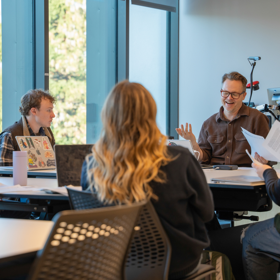 UP arts administration students engage in a class discussion with their professor at a table with laptops and documents; one gestures with papers, the other is viewed from behind.