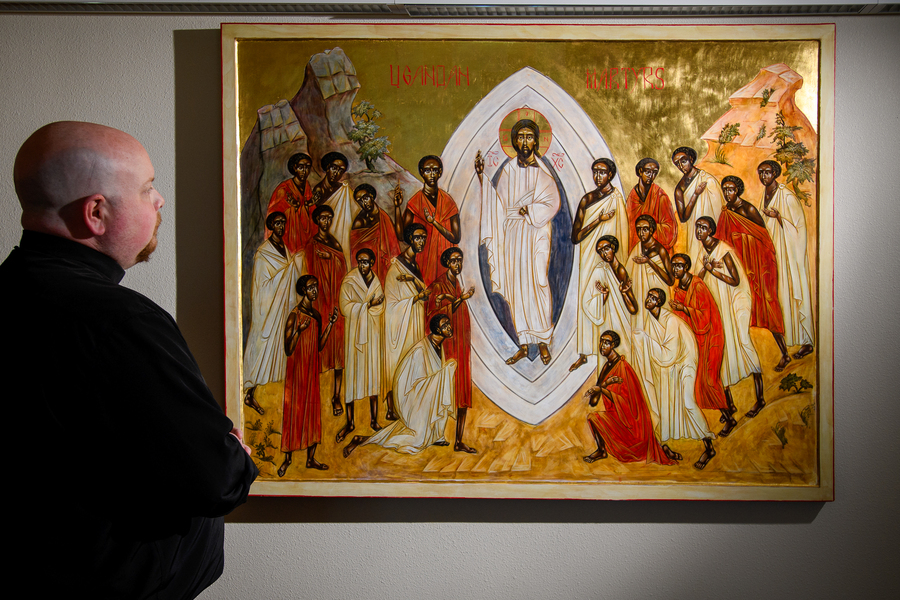 Father Tim Weed admires a Byzantine style painting depicting Jesus and a group of Ugandan martyrs. He stands to the side of the fully visible artwork with his back to the camera.