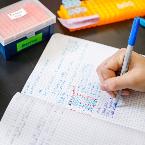 Close-up of a UP student's hand writing detailed notes on a grid-patterned notebook page with blue handwritten text, partially visible. 
