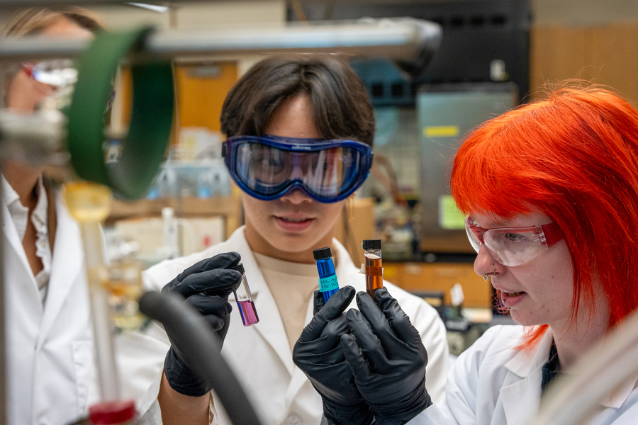 A person wearing safety goggles holds two vials, one filled with blue liquid and the other with orange liquid, in a laboratory setting.