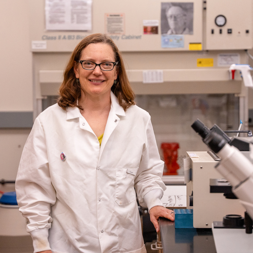 A University of Portland professor in a white lab coat smiles as she stands in a chemistry lab surrounded by instruments and equipment.