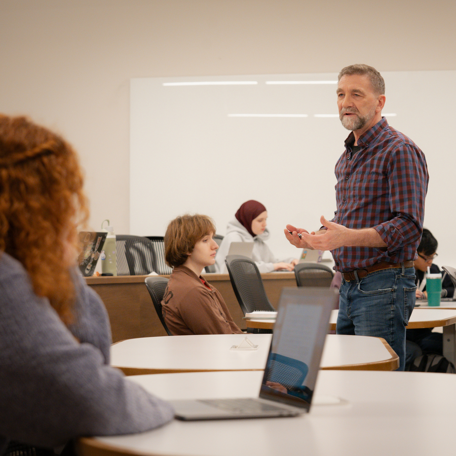 A UP Philosophy professor with short hair and a beard, wearing a plaid shirt, stands in a well-lit room with a plain white wall background lecturing students.