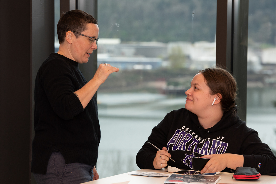 A professor speaks to a smiling student seated in a classroom with windows looking out onto the Willamette river in the background at the University of Portland.