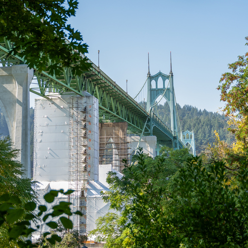 An view from below of the St Johns Bridge, a pale green suspension bridge with prominent towers and vertical cables, set against a clear sky, trees, and the hills of Forest Park in the background.