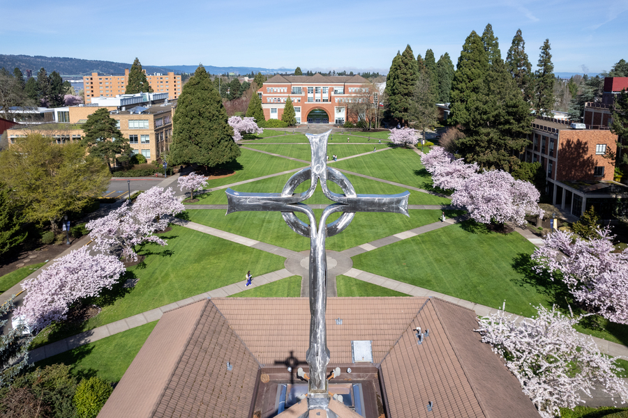 The UP bell tower cross is the primary focus of an aerial image with tall evergreens and pink blossomed cherry trees lining a verdant campus quad; hills and water are visible in the background under a clear sky.