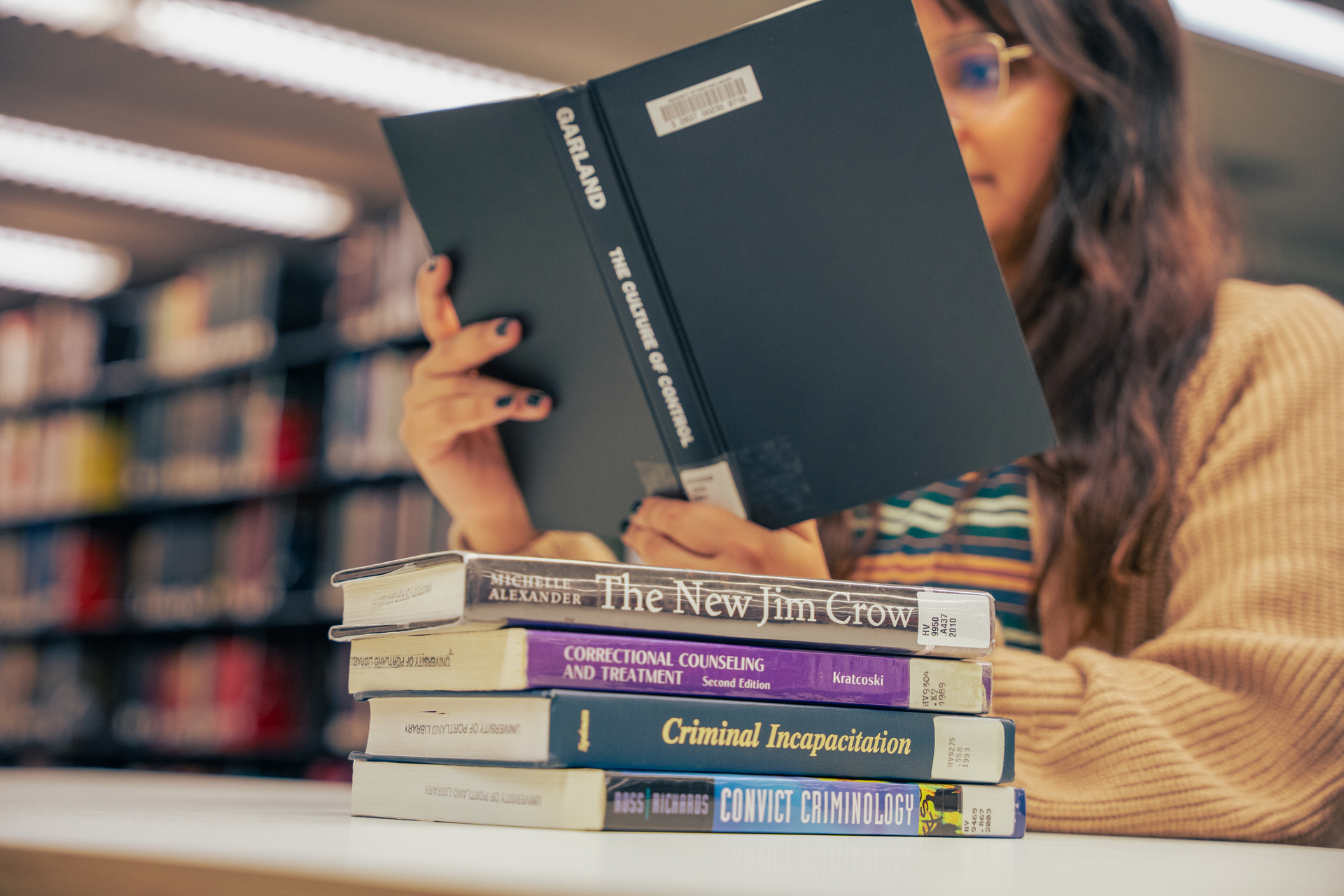 A low angle image of a student, seated at a table topped with a stack of books, their face partially obscured by the open book they are reading.