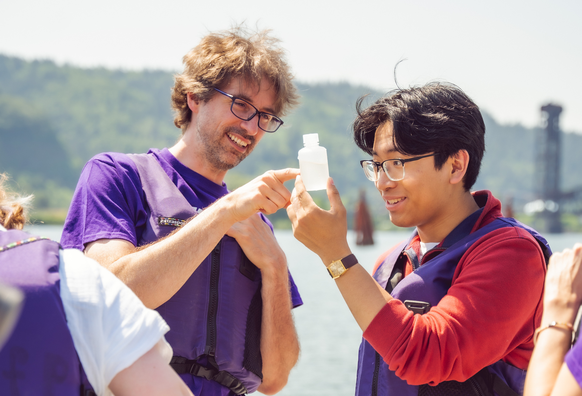 A UP biology professor and student smile while they examine a bottle of Willamette river water being sampled for an algae study on a sunny day with the river and hills in the background.