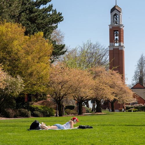 An University of Portland student reads a book while lying on a blanket outdoors on the Academic Quad, surrounded by grass, trees, and bushes on a sunny day. The Bell Tower is in the background. 