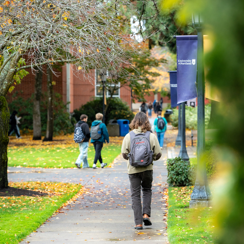 University of Portland students walk along the sidewalks of the academic quad, the ground is scattered with colorful fall leaves.