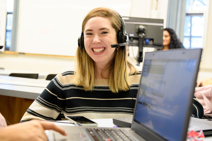 A UP student intern wearing a telephone headset smiles at the camera while working in the university relations office.