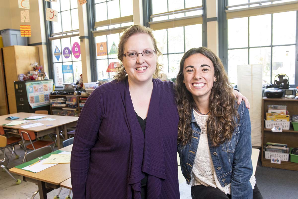 A University of Portland Professor of Education and an education student stand smiling together in a classroom with the professor’s arm around the student’s shoulder