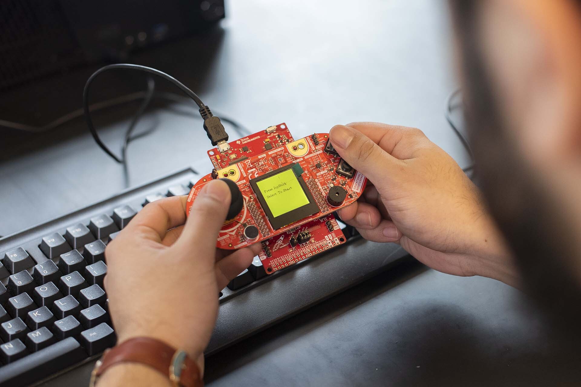 A close-up of a persons hands manipulating a circuit board, with part of the desk, keyboard, and monitor stand visible in the background.