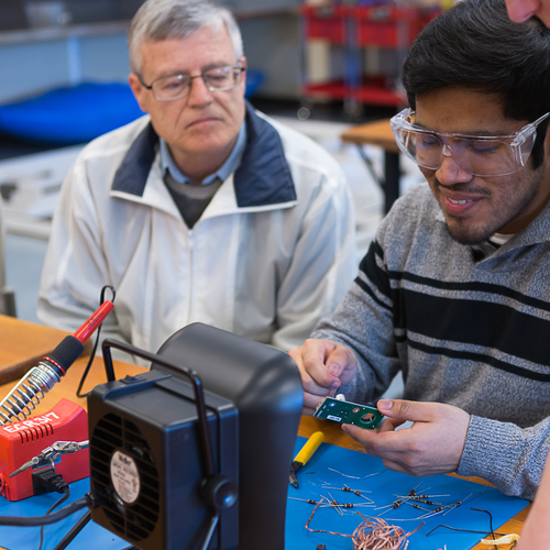 A close-up view of a professor seated at lab work station with an electrical engineering student. The professor watches as the smiling student in lab goggles manipulates a circuit.