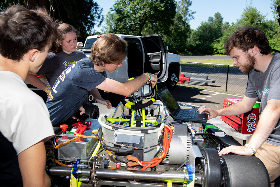 The UP racing team, a group of four students, lean over their electric go-kart to make adjustments to the equipment on a sunny day outdoors.