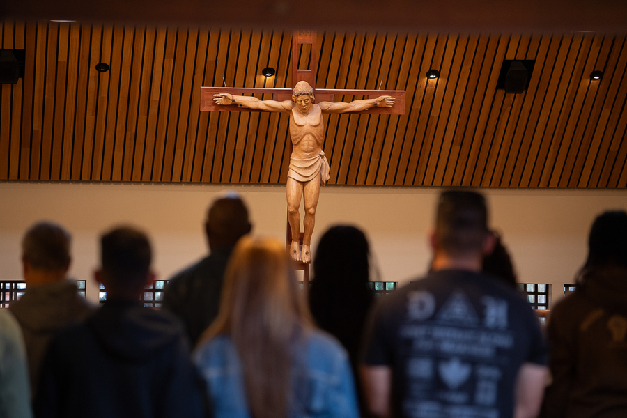 A hand carved wooden crucifix sculpture of Jesus in the Chapel of Christ the Teacher at the University of Portland faces the camera, with the backs of congregants silhouetted in the foreground.