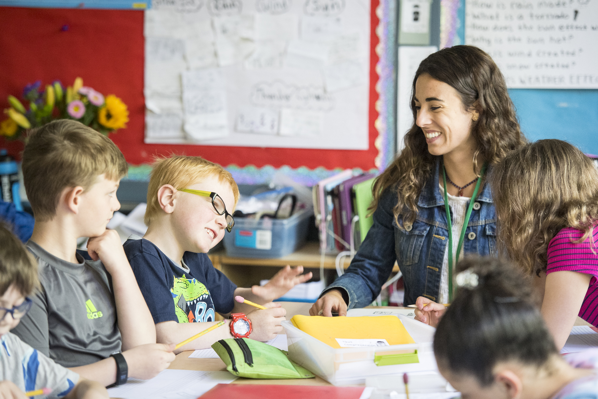 A University of Portland student in the elementary education programs sits at a round table with children in school classroom as they work with art supplies.