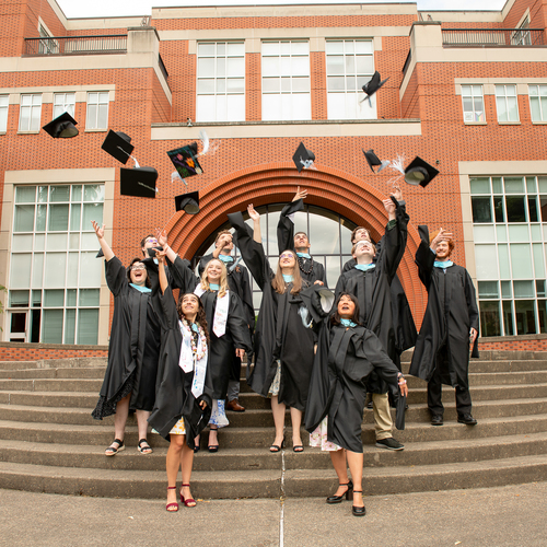 A group of University of Portland graduates from the Pacific Alliance for Catholic Education program, dressed in caps and gowns, stand on the steps of Franz Hall as they toss their mortar boards into the air in celebration.