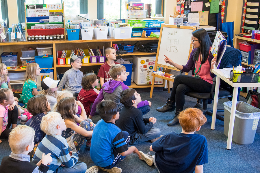 A University of Portland student in the elementary education programs sits in front of a group of children seated on the floor in school classroom as she reads aloud to them.