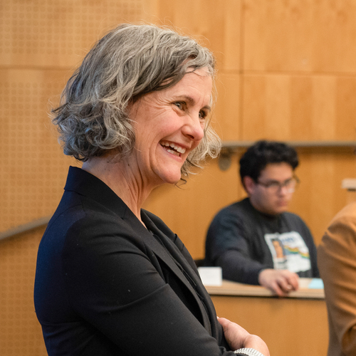 A close-up image of a University of Portland english professor smiling in the classroom during a lecture, a seated student is visible in the blurred background.