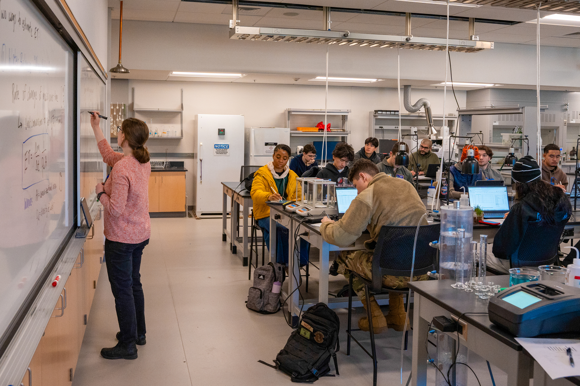 A University of Portland professor writes on a whiteboard filled with mathematical equations in a classroom, only their hand and marker visible. A light fixture and part of the ceiling appear in the background.