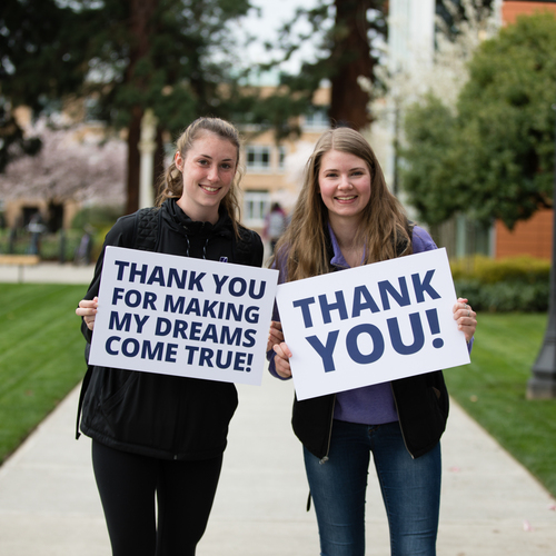 Two UP students with long brown hair smile for a posed picture holding signs that say thank you. Trees and a university building are in the background.
