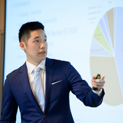 A UP business student, dressed in a blue suit and visible from shoulders to mid-chest, giving a presentation in front of projector screen displaying a pie chart.