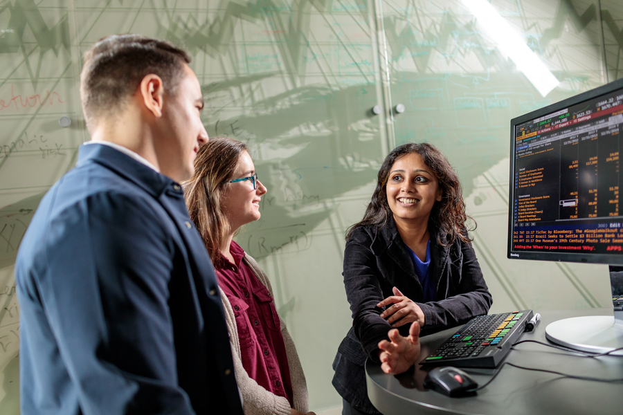 Two UP students stand smiling with their professor as they view the Bloomberg terminal during a business analytics class in Franz Hall.