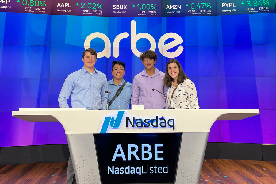 Four students from the UP Investment Association pose and smile from behind the NASDAQ podium at the Wall Street stock exchange in New York City.