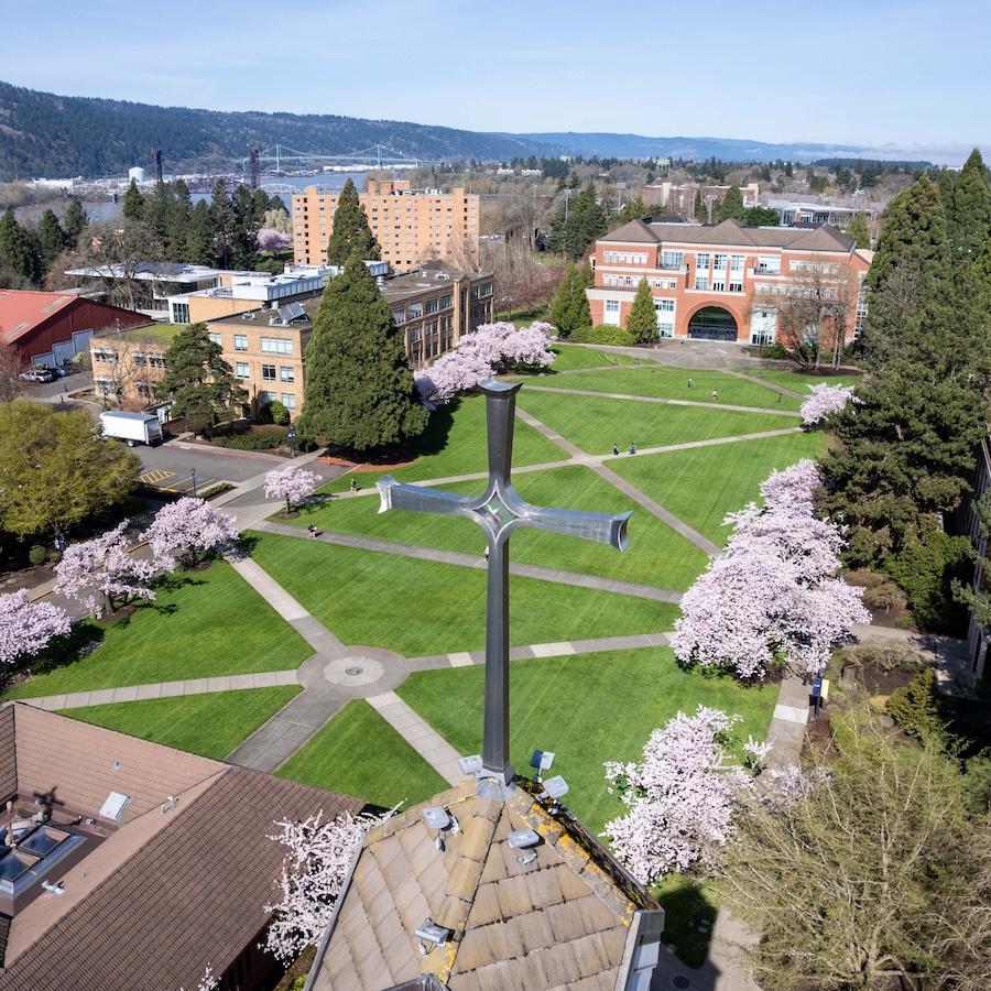 Sunny day aerial view of the cherry blossom-lined quad at University of Portland with the cross of the belltower in the forefront and Franz Hall in the distance